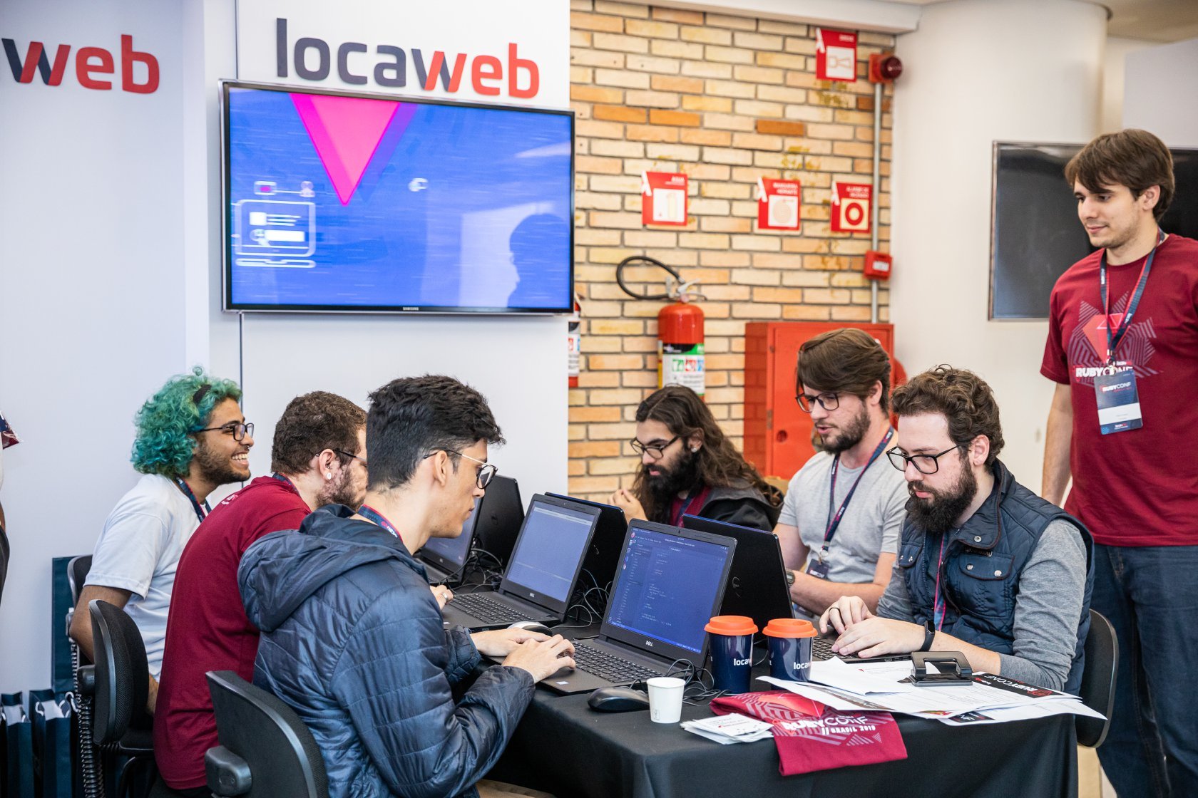 Escritório com pessoas em frente a seus computadores. Ao fundo, logo da Locaweb