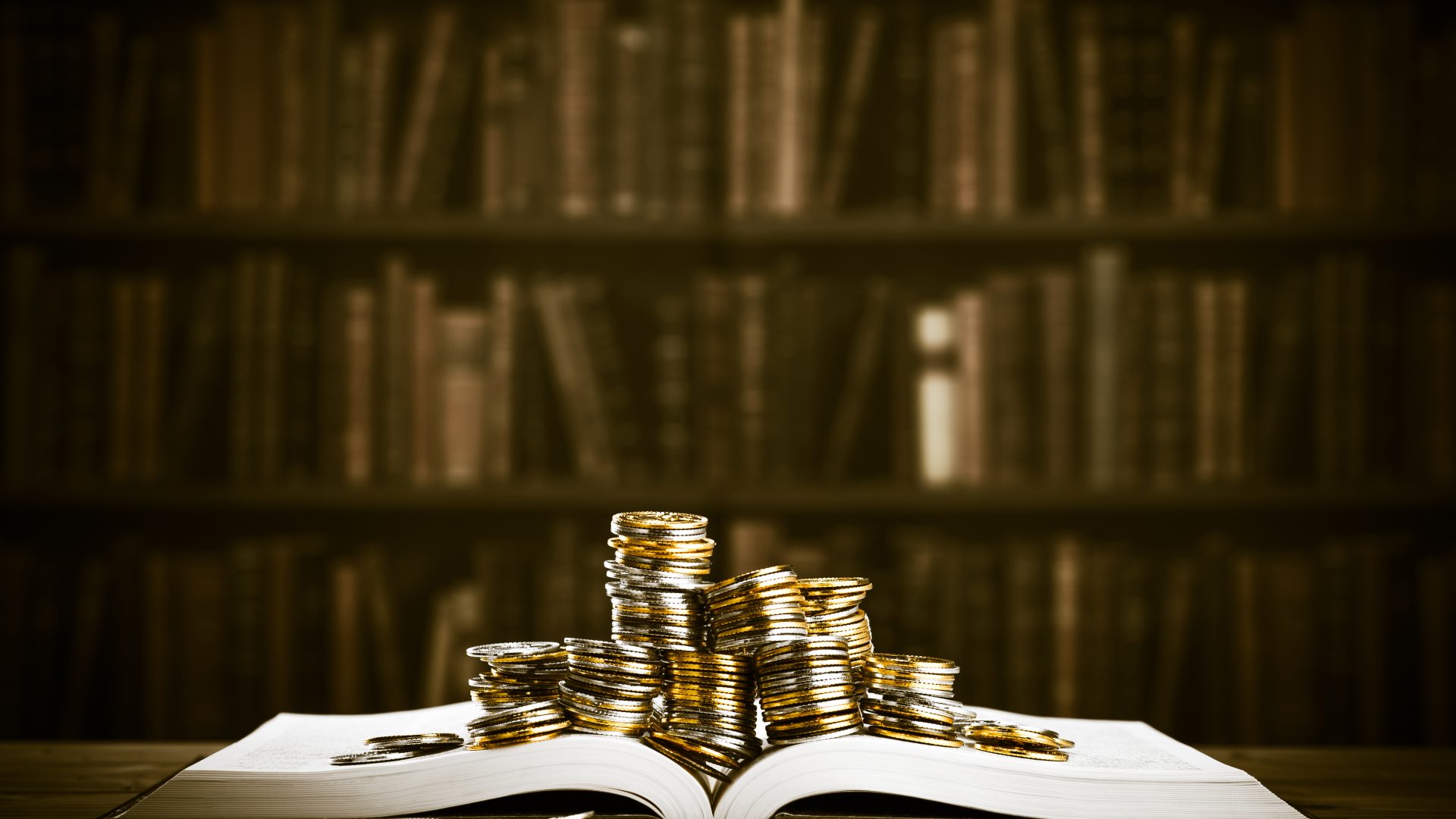 Moedas empilhadas em cima de um livro aberto. Ao fundo, uma estante.