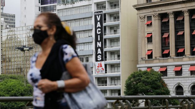 Mulher em primeiro plano do lado esquerdo da imagem caminha por uma rua de São Paulo. Ao fundo, um prédio com uma faixa na vertical com a frase "Vacinas Já"