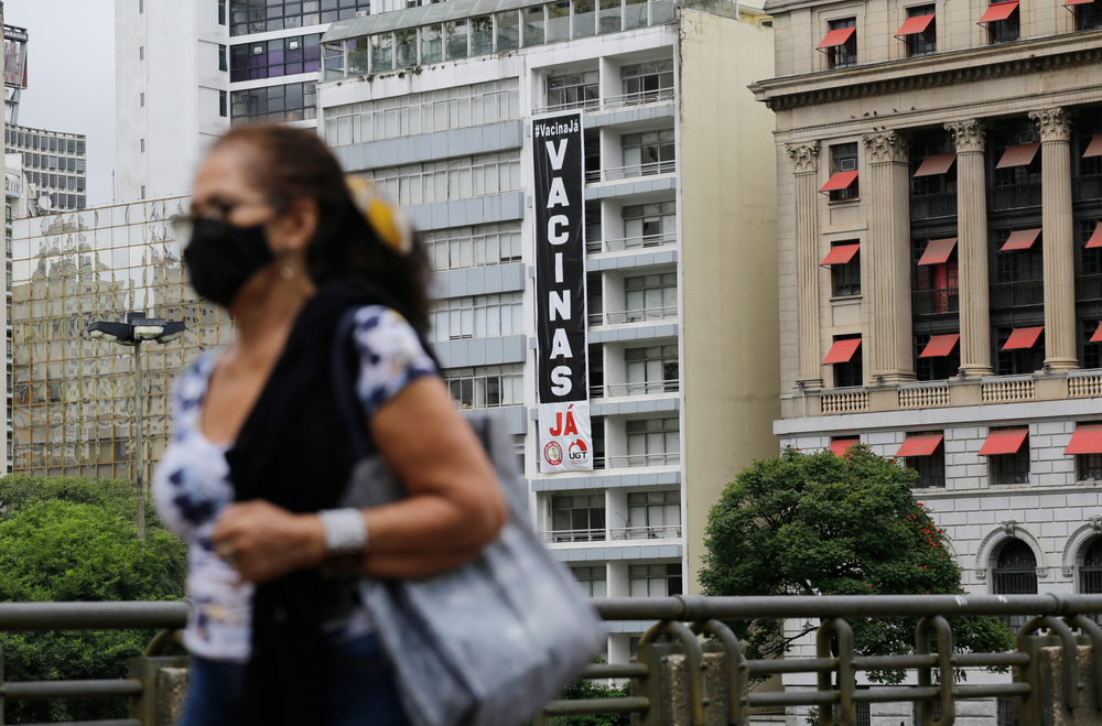 Mulher em primeiro plano do lado esquerdo da imagem caminha por uma rua de São Paulo. Ao fundo, um prédio com uma faixa na vertical com a frase "Vacinas Já"