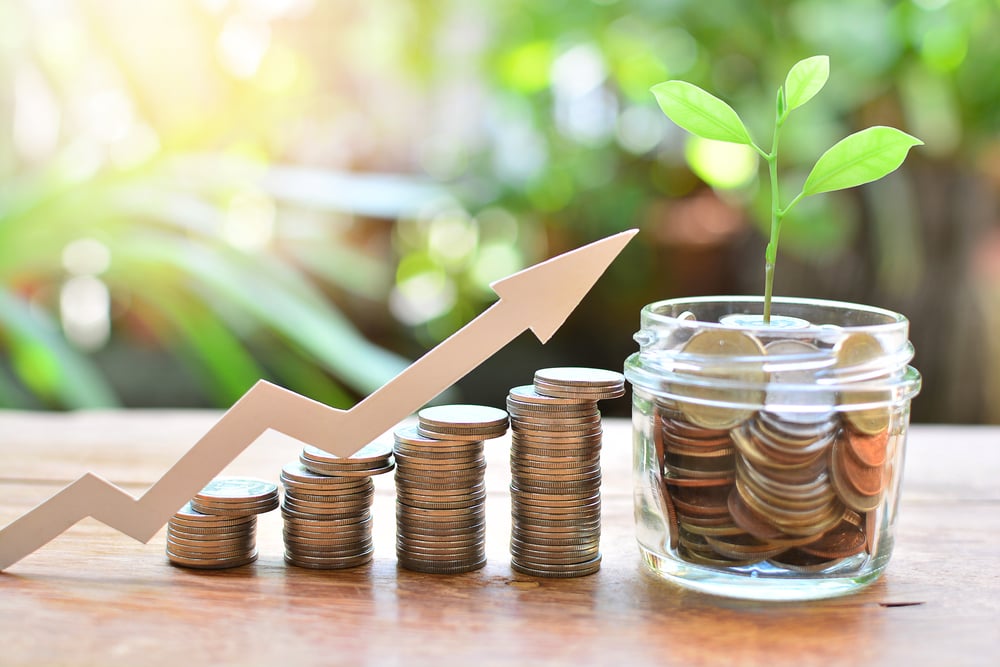 Moedas empilhadas,uma dos lado da outra, em tamanho ascendente, com montagem de seta indicando crescimento e um pote com moedas ao final (direita), simbolizando investimentos lucrativos