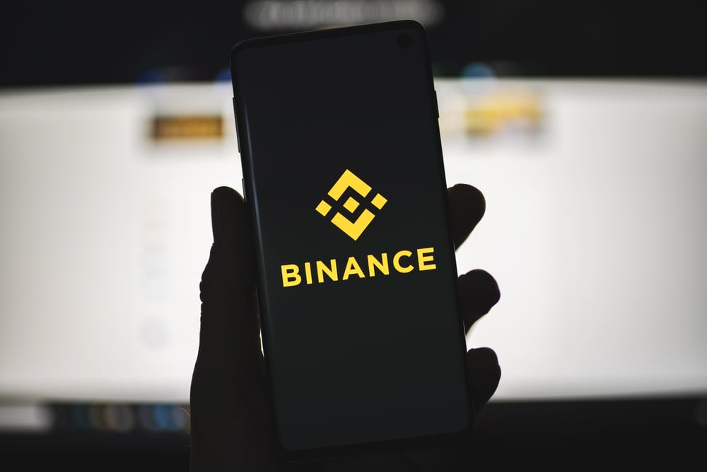 Uma mão segura um smartphone com a tela preta e o logo da corretora Binance em amarelo no centro