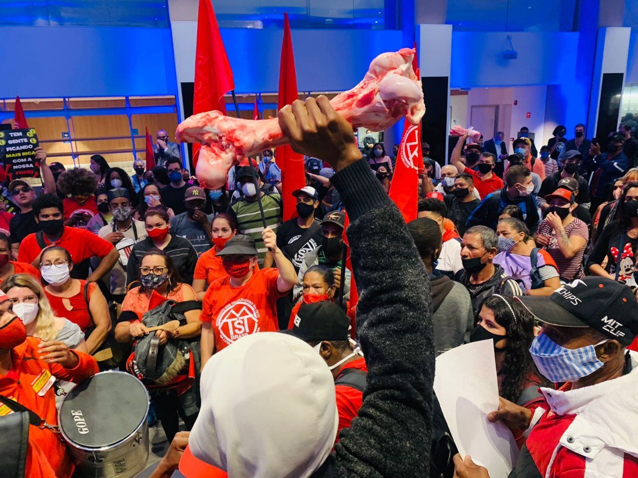 Integrante do MTST segurando um osso bovino em primeiro plano, com outros manifestantes vestidos de vermelho ocupando a sede da B3