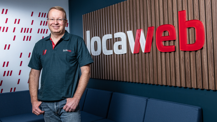 Foto de Fernando Cirne, CEO da Locaweb (LWSA3). Ele está de pé, usando calça jeans e camisa preta; ao fundo, um sofá azul e uma parede com revestimento em madeira e o logo da Locaweb