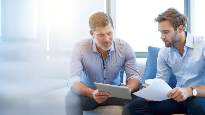 Dois homens brancos de terno conversando; um com um tablet na mão, outro com papéis na mão, representando soft skills