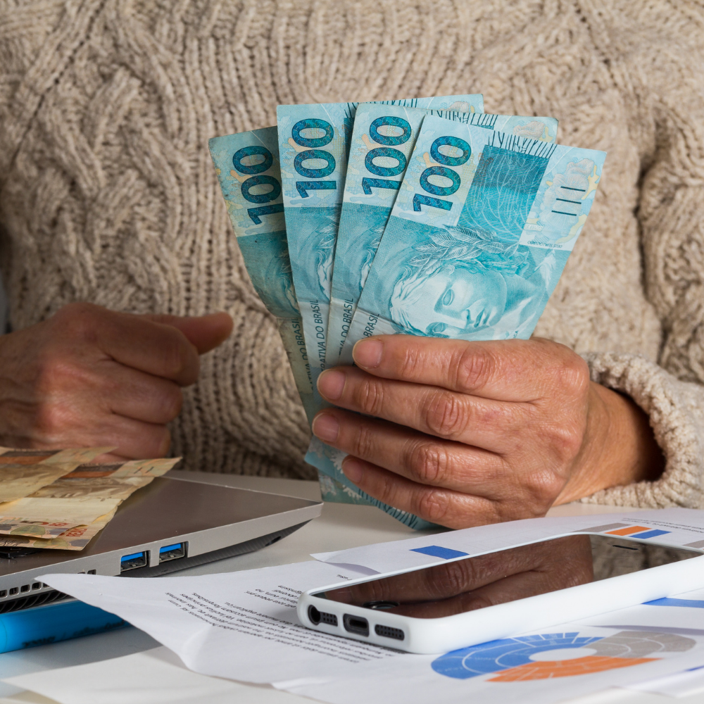 Uma pessoa segura notas de 100 reais tendo um celular e papéis sobre a mesa que está a sua frente