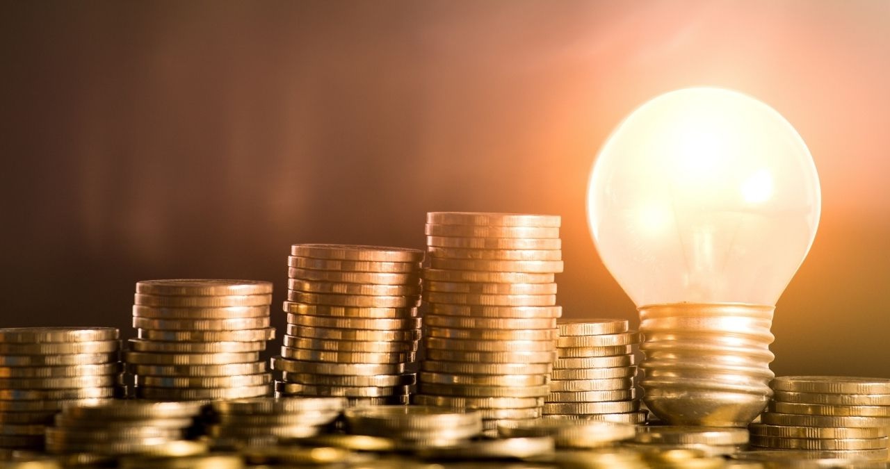 Imagem mostrando pilhas de moedas cada vez maiores, com uma lâmpada acesa à direita. Simboliza a alta da Selic e dos juros e as ideias de investimento em renda fixa e renda variável nesse cenário
