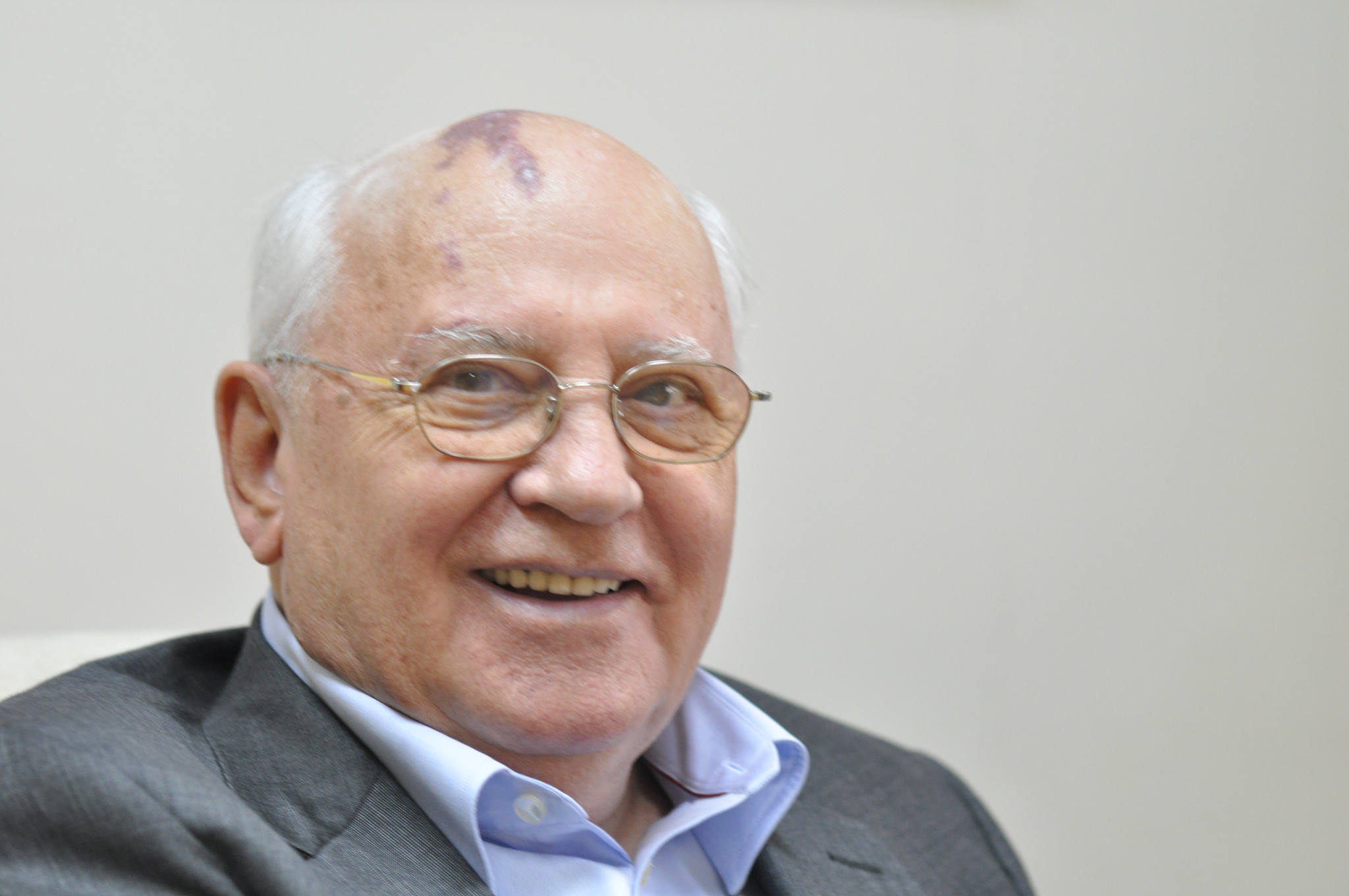Foto de meio corpo de Mikhail Gorbachev, usando óculos e sorrindo, vestindo terno cinza e camisa azul