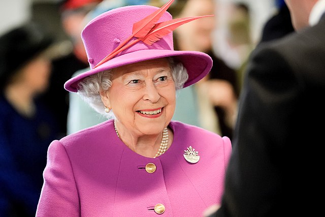 Foto de meio corpo da rainha Elizabeth II, vestindo roupas roxas e de chapéu com pena