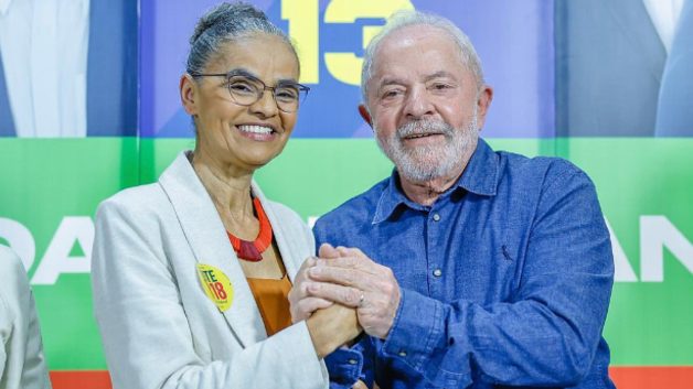 Marina Silva veste blazer claro, blusa mostarda e colar colorido. Ela dá as mãos para Lula, que está de camisa jeans. Ao fundo um painel da campanha eleitoral de Lula em 2022.