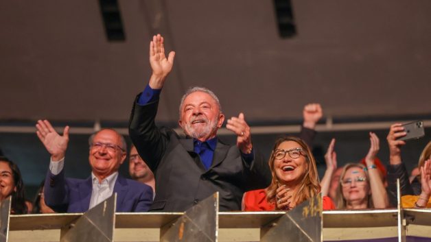 Lula em pé, em palanque, batendo palmas. Ao lado dele, Geraldo Alckmin dá tchau e do outro lado, a esposa de Lula, Janja, aplaude