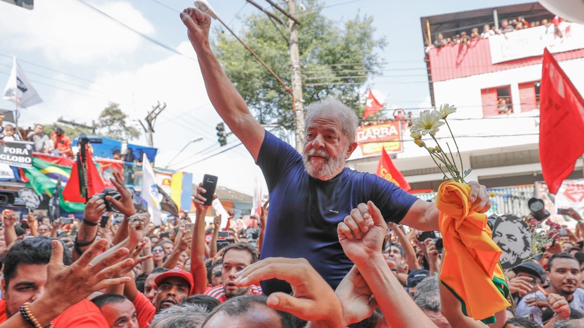 Lula veste camiseta azul marinho, segura uma camiseta laranja e flores brancas em uma mão, a outra está erguida. Ele está nos braços no povo, no meio de uma multidão