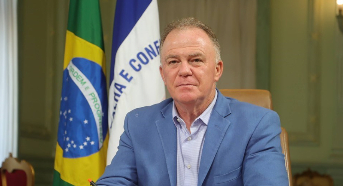 Homem vestido de blazer azul e camisa azula mais clara com bandeira do Brasil e do ES atrás e um óculos de grau sobre a mesa