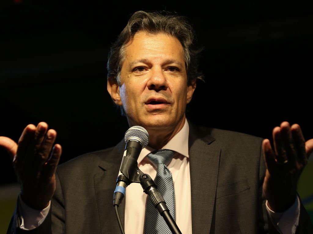 Fernando Haddad fala em microfone, com os braços abertos. Ele veste terno cinza, camisa clara e gravata azul.