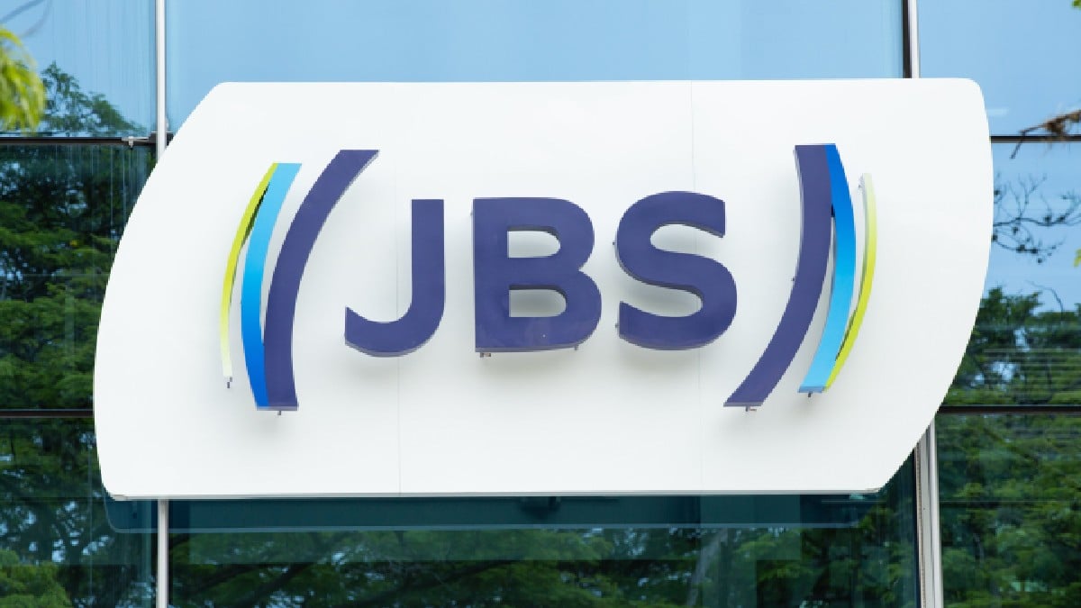 Logo da companhia JBS, ao fundo céu azul e árvores espelhados na fachada do prédio