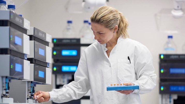 A imagem mostra uma farmacêutica loira, com jaleco branco, em um laboratório cheio de equipamentos. Ela segura um suporte com várias amostras. Ozempic