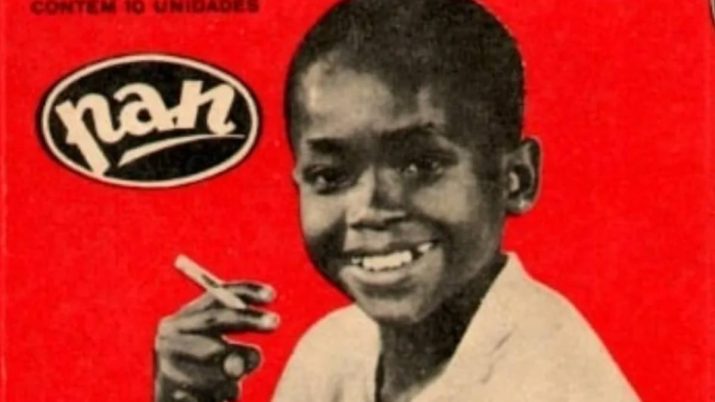 Foto antiga em fundo vermelho, com um garoto preto segurando um cigarro de chocolate e a marca Pan ao lado, em preto