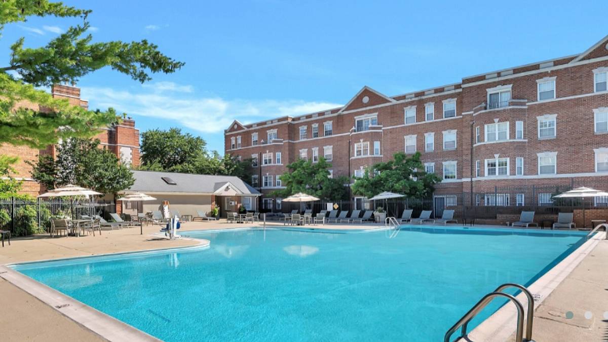 Foto de uma piscina grande, com cadeiras de praia em volta, e um prédio de tijolinhos ao fundo. Faz sol e árvores também fazem parte da paisagem.