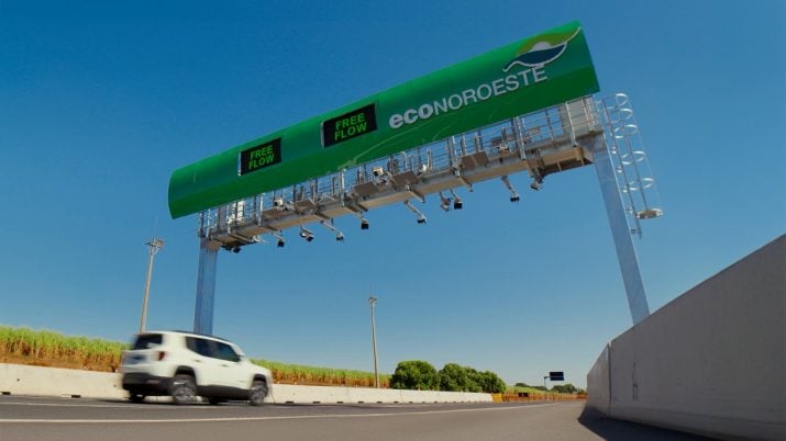 Rodovia vista da lateral, de cima para baixo, com uma placa em cima, com os dizeres "EcoNoroeste" em branco e logo da EcoRodovias acima. Há um carro branco passando na pista.