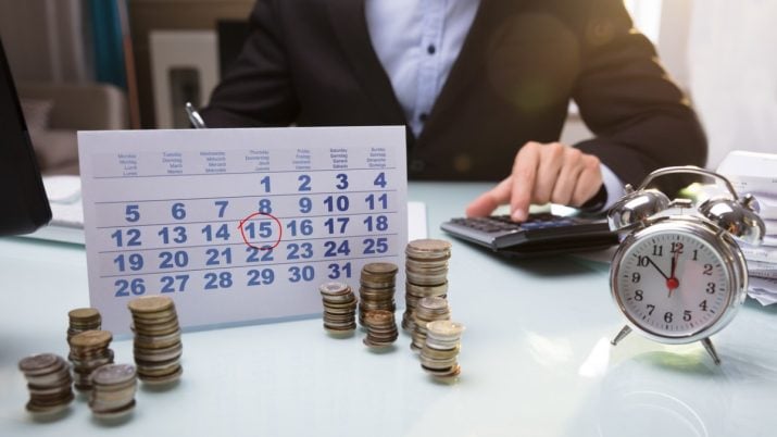 Imagem de calendário com pilhas de moedas na frente e, atás, um homem de terno usando uma calculadora