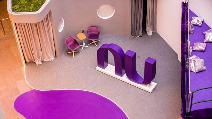 Espaço do Nubank com cadeiras na lateral esquerda e travesseiros na lateral direita. No centro, símbolo do Nubank