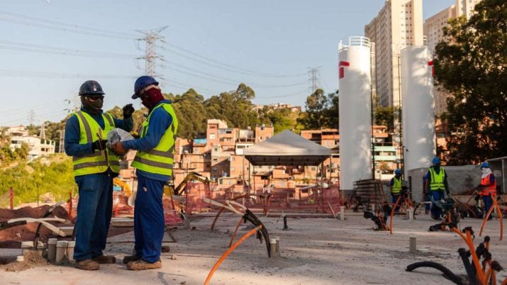 Dois técnicos de construção conversando na lateral esquerda. Atrás, há duas torres brancas com símbolo vermelho da Tenda, casas e prédios.