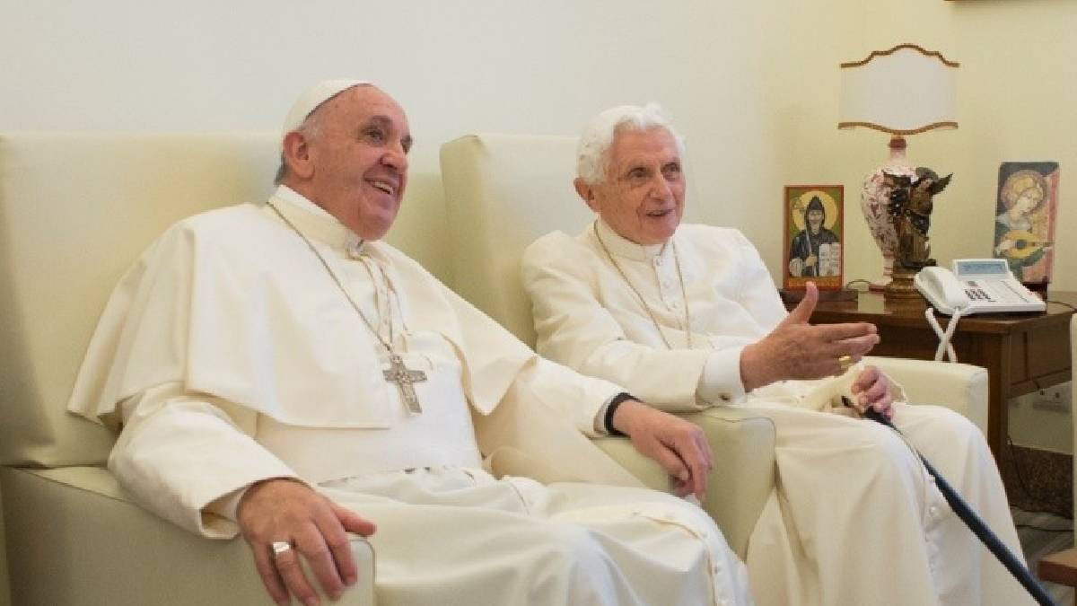 Imagem do papa Francisco, vestido de branco, sentado em uma poltrona branca. Ao seu lado, Bento 16, que também já foi papa.